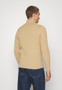 Suéter de punto cableado beige con cuello alto, puños acanalados y dobladillo. La textura de la tela presenta un patrón en relieve para añadir dimensión.