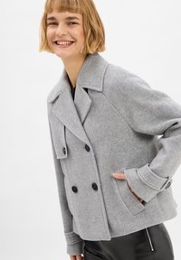 Grijze dubbelbreasted peacoat van wolmix, met grote zwarte knopen, brede revers en geribde manchetten. Model poseert met handen in de zakken.