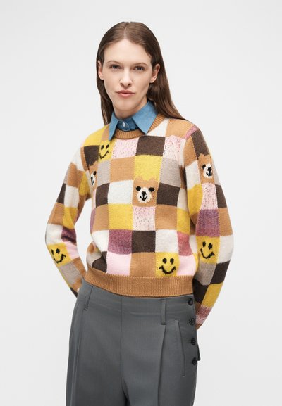 Femme portant un pull à carreaux avec des carrés jaunes, marron et roses ornés de visages souriants et de têtes d'ours, associé à un pantalon gris.