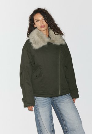 Femme aux cheveux bouclés portant une veste vert foncé avec un grand col en fourrure gris clair et un jean bleu clair sur un fond uni.