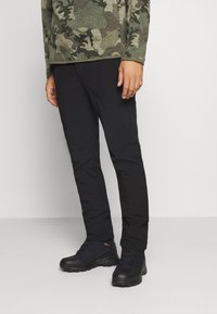 Pantalon noir à coupe slim, accompagné de bottes noires robustes. La chemise arbore un motif camouflage dans des tons de vert et de marron.