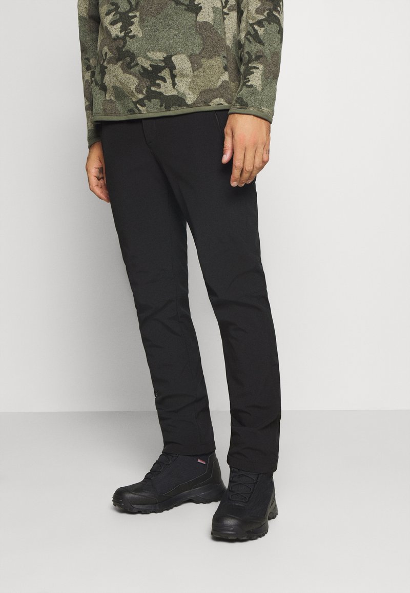 Pantalon noir à coupe slim, accompagné de bottes noires robustes. La chemise arbore un motif camouflage dans des tons de vert et de marron.