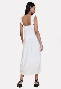Abito maxi bianco a strati con spalline a volant e schiena quadrata. Realizzato in tessuto leggero, presenta dettagli sulle cuciture.