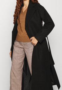 Long black wool coat with shawl collar, open front, and large armholes over a brown sweater and plaid trousers.