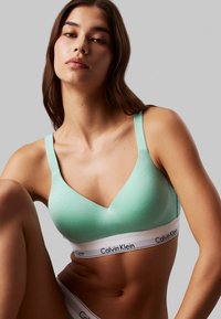 Bralette v svetlozeleni barvi, izdelan iz gladkega materiala, z obliko v-izreza; ima belo elastično trak, natisnjen z "Calvin Klein."