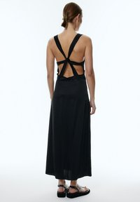 Zwarte maxidress met open rug, met knoopbandjes en een vloeiende silhouet. Gemaakt van een zachte stof met een subtiele glans.