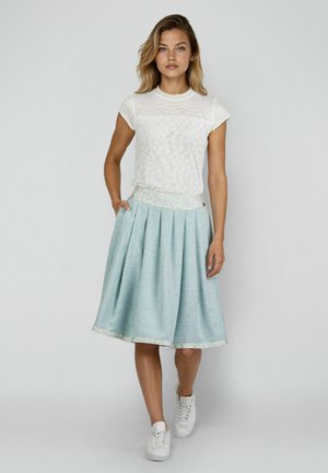 Junge Frau, die ein weißes Spitzenoberteil mit kurzen Ärmeln, einen hellblauen Faltenrock und weiße Turnschuhe trägt, vor einem schlichten grauen Hintergrund steht.