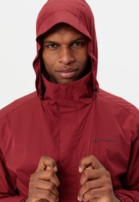 Červená voděodolná bunda s vysokým límcem a nastavitelnou kapucí. Má zapínání na zip v přední části a název značky "Vaude" na hrudi.