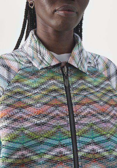 Veste en tricot avec un col haut, fermeture éclair sur le devant et motif zigzag multicolore dans des textures vertes, oranges, violettes et noires.