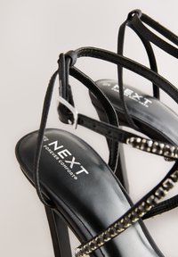 Črne sandale z visokimi petami in trakovi, s sijočim zaključkom, okrašene z dekorativnim kovinskim poudarkom in oblazinjenimi vložki z napisom "NEXT Forever Comfort".