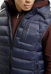 Mężczyzna w granatowej, pikowanej kamizelce noszący ją na bordowej bluzie z zamkiem, prezentujący zamykane na zamek kieszenie kamizelki oraz logo "CROSS JEANS" na piersi.