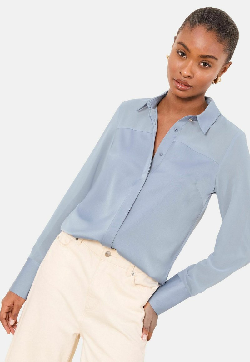 Gaiši zilas, garām piedurknēm pogājamas krekliņš ar mīkstu tekstūru, klasisku apkakli un priekšējo yoke šuvi, kombinēts ar krēmkrāsas biksēm.