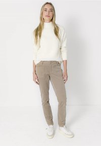 Blondýnka stojící, oblečená v bílém texturovaném svetru, béžových kalhotách a bílých teniskách, na jednobarevném bílém pozadí.