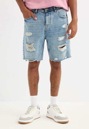 Hombre con pantalones cortos de mezclilla azul claro desgastados, calcetines blancos y zapatillas blancas, de pie con una mano en el bolsillo.