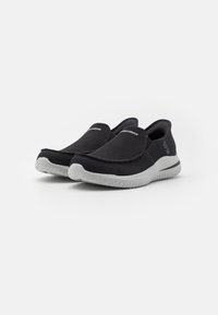 Svarte slip-on sko med en teksturert mesh-overdel og en lett hvit gummi-såle. Har minimal merkevare og et strømlinjeformet design.