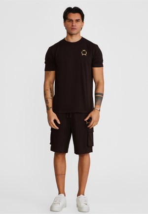 Hombre con pelo oscuro que lleva una camiseta negra y pantalones cortos cargo con un emblema dorado, zapatillas blancas, de pie frente a un fondo gris liso.