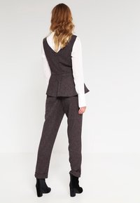 Chaleco de tweed marrón con diseño de peplum, combinado con pantalones ajustados a juego. Blusa de manga larga blanca debajo. Botines de tacón negro.