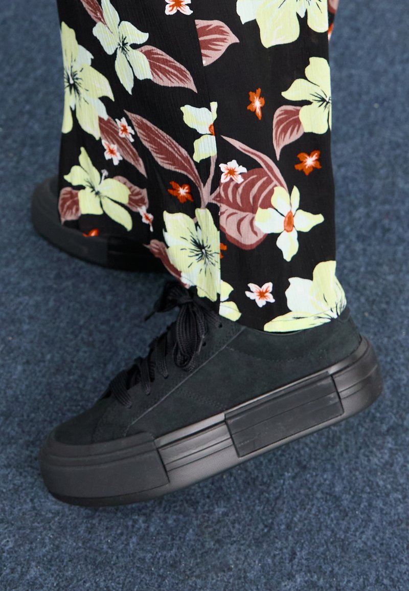 Schwarze Wildleder-Sneaker mit einer dicken schwarzen Gummisohle und Schnürungen. Der Hintergrund zeigt einen blumigen Stoff mit lebhaften Farben.