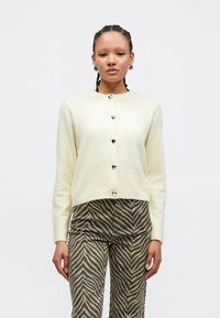 Cardigan crème à encolure ronde avec des boutons dorés en forme de cœur, assorti à un pantalon marron à motif zébré.