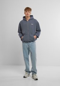 Gray zip-up hoodie with a front pocket and embroidered logo, paired with light blue jeans and silver sneakers. Smooth texture and relaxed fit.