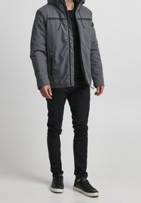 Grå isolerad jacka med huva, kontrasterande svarta dragkedjor och två sidofickor. Kombinerad med svarta byxor och svarta sneakers.