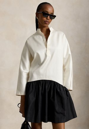 Femme portant des lunettes de soleil noires et un haut blanc à manches longues avec une jupe noire froncée, tenant un petit sac à main noir.