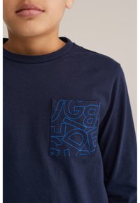 Námornícka tričko s dlhým rukávom, okrúhlym výstrihom a modrou vzorovanou náprsnou vreckom s výraznými písmenami. Materiál je mäkký a bavlnený.