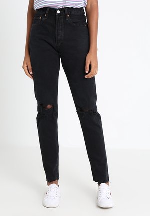 Modelka w wysokich, czarnych, podartych dżinsach z surowymi brzegami, w połączeniu z paskowaną koszulą i białymi trampkami na jednolitym białym tle.