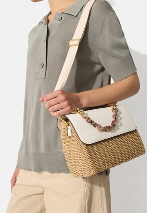 Persoon in een lichtgrijs polo met korte mouwen en beige broek houdt een geweven strooien handtas vast met een witte flap en gemarmerde kettingdetails.