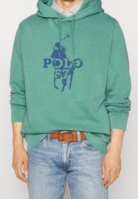Grüne Baumwollkapuzenjacke mit einem großen navyfarbenen "POLO 67"-Grafik und einem Reiterdruck. Getragen über einem grauen Hemd mit Kragen und hellblauen Jeans.