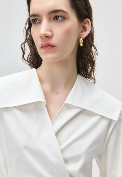 Młoda kobieta z kręconymi brązowymi włosami, ubrana w białą bluzkę z kołnierzykiem i złote kolczyki koła, patrząca lekko na bok z neutralnym wyrazem twarzy.