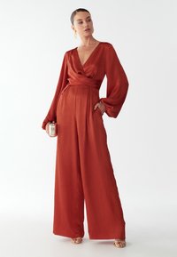 Rudos spalvos kombinezonas su ilgomis rankovėmis, giliu V formos iškirpimu ir surištu juosmeniu. Pagamintas iš lygaus audinio, turi plačius kelnes.