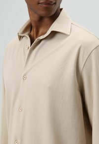 Beige, overhemd met lange mouwen en knopen, met een kraag, voorzien van een gladde textuur en twee zichtbare knopen. Eenvoudig ontwerp, minimale details.