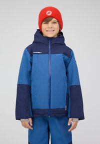 Veste pour enfants bleu et marine avec fermeture éclair, capuche et logo. Texture lisse avec un design contrasté. Associée à un bonnet rouge en tricot.