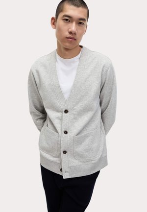Světlé šedé kardigan vyrobený z měkké tkaniny, s výstřihem do V, zapínáním na knoflíky, dvěma předními kapsami a žebrovaným lemem.