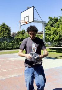 Een persoon in een zwart T-shirt met een cartoonbeer houdt een bundel zwarte spullen vast op een basketbalveld, met groenigheid en een ring in het zicht.