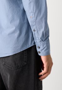 Light blue long-sleeve shirt with buttoned cuffs; features contrasting buttons and orange stitching detail; paired with dark gray pants.