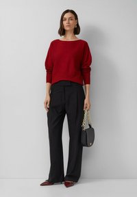 Roter Strickpullover mit geripptem Saum, kombiniert mit schwarzen Hosen. Es gibt eine schwarze Handtasche mit Kettenriemen und burgunderfarbene spitze Schuhe.