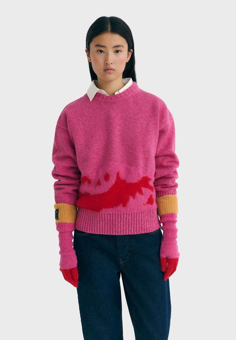 Ung kvinde iført en sweater med lyserøde og røde mønstre, der matcher hendes handsker, samt mørke jeans, står op ad en ensfarvet baggrund.