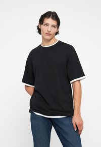 Camiseta de algodón negra con cuello redondo y ribetes de manga en blanco, con un corte relajado y mangas cortas, combinada con jeans de mezclilla azules.