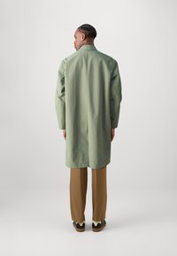 Lungo cappotto verde in tessuto morbido, con taglio dritto, una spacco sul retro, abbinato a pantaloni marroni e scarpe nere.