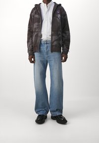 Chaqueta con capucha de tie-dye marrón, camisa blanca de botones, jeans de pierna ancha azul claro y calzado negro. Gráfico sutil en la chaqueta.
