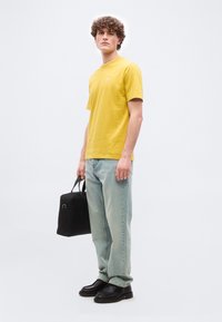 Νεαρός άνδρας με καστανά σγουρά μαλλιά, που φοράει κίτρινη φανέλα, ανοιχτό μπλε τζιν, μαύρα παπούτσια, κρατώντας μια μαύρη ορθογώνια τσάντα δίπλα του.