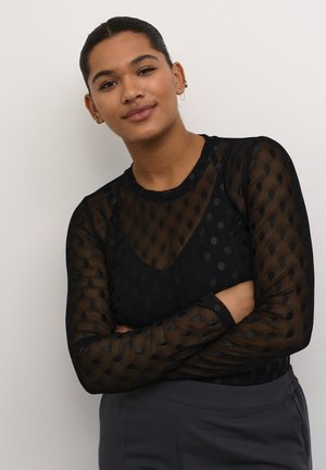 Zwarte mesh top met lange mouwen, voorzien van een stippenpatroon, semi-transparante textuur en een rond decolleté. Gecombineerd met grijze high-waisted broek.