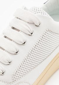 Baskets blanches en cuir, avec un design perforé sur le côté, des lacets blancs texturés et une semelle en caoutchouc beige.