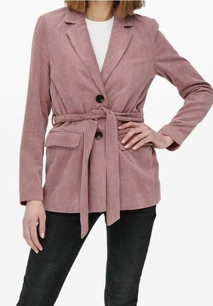 Femme portant un blazer en velours côtelé rose poussiéreux avec des boutons noirs, une ceinture à la taille, des poches à rabat, une chemise blanche et un pantalon noir.