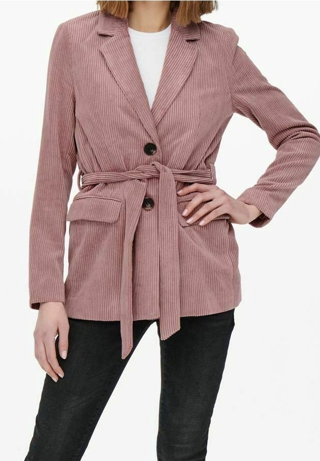 Femme portant un blazer en velours côtelé rose poussiéreux avec des boutons noirs, une ceinture à la taille, des poches à rabat, une chemise blanche et un pantalon noir.