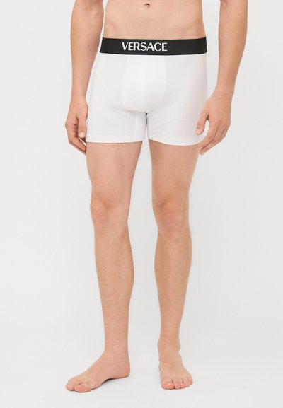 Weiße Boxershorts mit einem schwarzen Bund, auf dem das "VERSACE"-Logo zu sehen ist. Glattes Material, eng anliegendes Design und mittellange Beine.