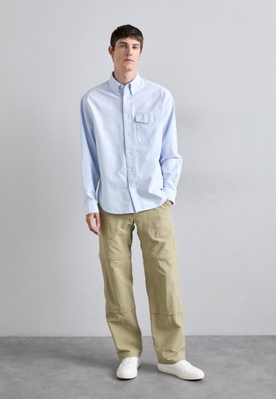 Chemise boutonnée bleu clair avec une poche poitrine, associée à un pantalon cargo beige avec de grandes poches et une coupe décontractée. Des baskets blanches complètent le look.