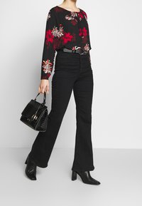 Blusa negra con estampado floral, de mangas largas y escote fruncido; combinada con unos pantalones vaqueros de tiro alto, negros y acampanados, y botas de tacón negras. Sosteniendo un bolso negro.